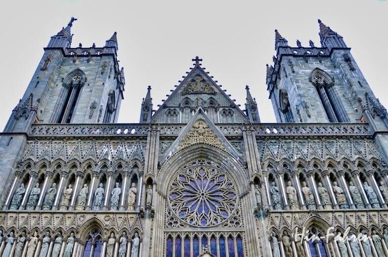 Fassade des Nidarosdoms: Eine Nahaufnahme der Fassade einer historischen Steinkathedrale in Trondheim, aufgenommen vom Wohnmobil aus, mit zwei hohen Türmen, Bogenfenstern, kunstvollen Schnitzereien und einer großen runden Glasmalerei in Form einer Rosette. Der Himmel darüber ist klar und hellgrau.