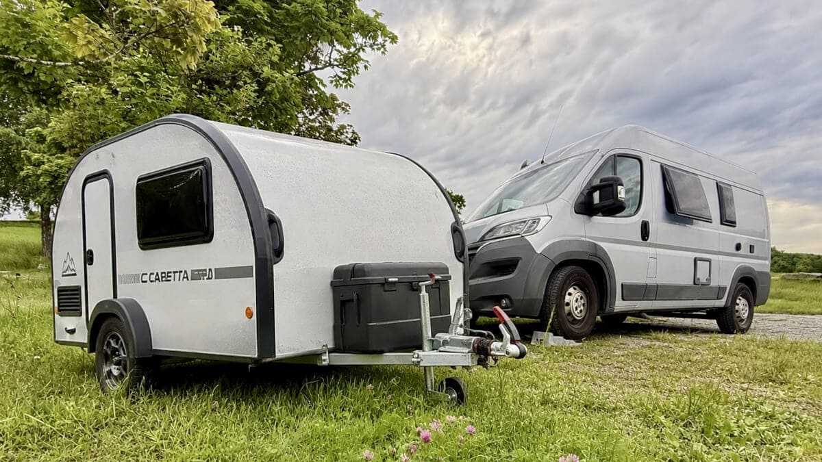 Unter einem bewölkten Himmel steht ein kompakter silberner Wohnwagen auf einer Wiese neben einem größeren silbernen Wohnmobil, im Hintergrund sind grüne Bäume zu sehen.