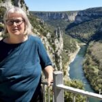 Eine Person (Katja Hein) mit kurzen grauen Haaren und Brille steht an einem Metallgeländer und blickt an einem sonnigen Tag auf einen malerischen Fluss, der sich durch eine tiefe, grüne Schlucht schlängelt.