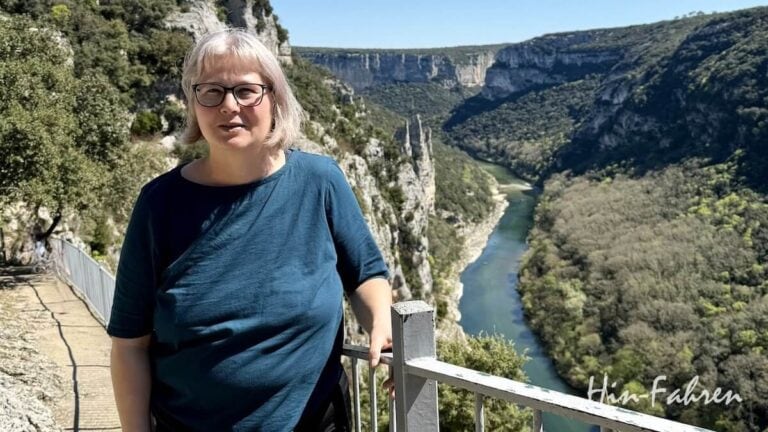 Eine Person (Katja Hein) mit kurzen grauen Haaren und Brille steht an einem Metallgeländer und blickt an einem sonnigen Tag auf einen malerischen Fluss, der sich durch eine tiefe, grüne Schlucht schlängelt.