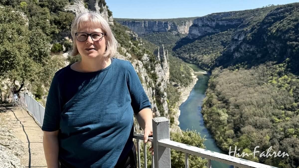 Eine Person (Katja Hein) mit kurzen grauen Haaren und Brille steht an einem Metallgeländer und blickt an einem sonnigen Tag auf einen malerischen Fluss, der sich durch eine tiefe, grüne Schlucht schlängelt.
