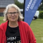 Katjas Campingtipps: Eine lächelnde Frau mit grauen Haaren und Brille steht im Freien und trägt eine rote Jacke und ein schwarzes T-Shirt. Hinter ihr sind eine blaue „CARAVAN DÜSSELDORF“-Flagge, Grünpflanzen, ein weißes Wohnmobil und „Hin-Fahren“ zu sehen – die perfekte Inspiration für Camping- und Wohnmobiltipps.