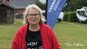 Katjas Campingtipps: Eine lächelnde Frau mit grauen Haaren und Brille steht im Freien und trägt eine rote Jacke und ein schwarzes T-Shirt. Hinter ihr sind eine blaue „CARAVAN DÜSSELDORF“-Flagge, Grünpflanzen, ein weißes Wohnmobil und „Hin-Fahren“ zu sehen – die perfekte Inspiration für Camping- und Wohnmobiltipps.
