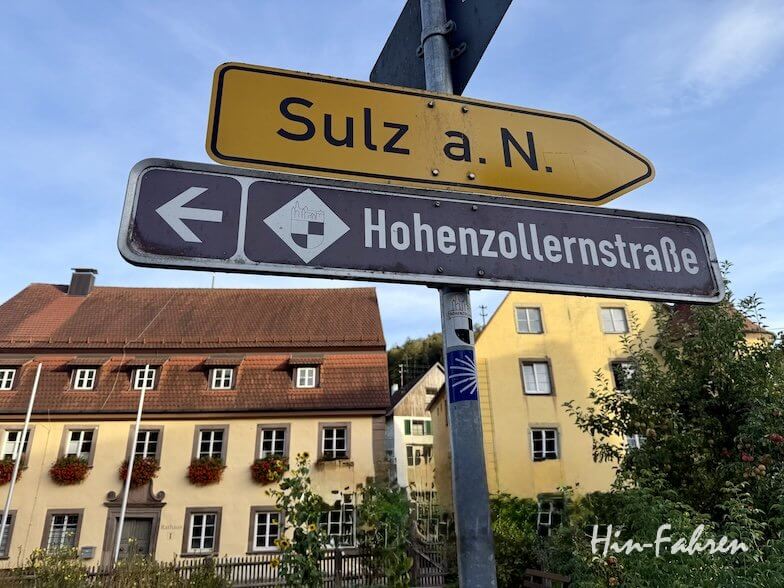 An einer Straßenecke stehen zwei Straßenschilder: Das obere gelbe Schild weist den Weg nach Sulz a. N., während das untere braune Schild den Wohnmobilverkehr nach links in die Hohenzollernstraße leitet. Im Hintergrund sind traditionelle deutsche Gebäude mit roten Dächern und Blumenkästen zu sehen.