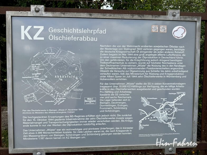 Eine Informationstafel im Freien in der Hohenzollernstraße mit dem Titel KZ-Geschichtslehrpfad Ölschieferabbau, die zwei historische Karten und einen deutschen Text enthält, in dem das Zwangsarbeitsprojekt Unternehmen Wüste im Bergbau während des Zweiten Weltkriegs beschrieben wird.