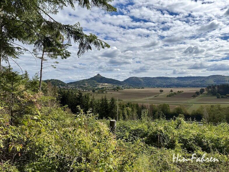 Ein Blick von einem bewaldeten Hügel entlang der Hohenzollernstraße auf ein weites, flaches Tal mit einer Burg auf einem entfernten Berggipfel.