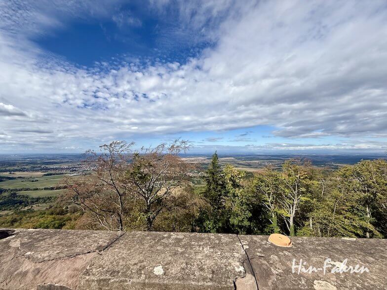 Panoramablick auf eine Landschaft mit Bäumen, Feldern und Ortschaften, aufgenommen von einem Aussichtspunkt aus Stein an der Hohenzollernstraße unter einem bewölkten blauen Himmel.