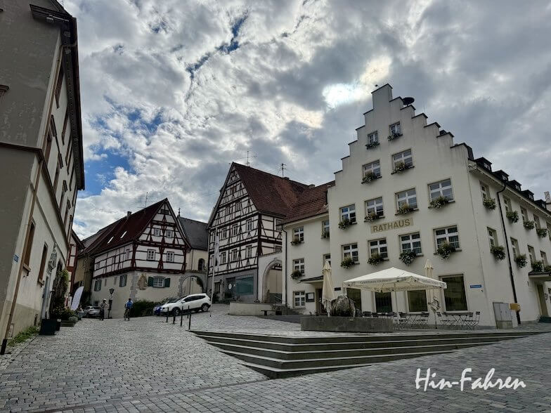 Ein mit Kopfsteinpflaster gepflasterter Stadtplatz in der Nähe der Hohenzollernstraße mit einem weißen Rathausgebäude mit Blumenkästen und einem historischen Fachwerkhaus unter bewölktem Himmel.