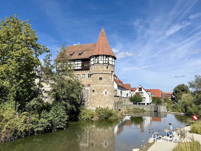 Ein mittelalterlicher Steinturm mit kegelförmigem Dach steht an einem Fluss in einer Stadt mit Fachwerkhäusern in der Nähe des Wohnmobilstellplatzes an der Hohenzollernstraße.
