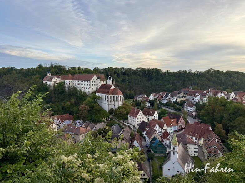 Blick von oben auf eine Stadt in der Nähe der Hohenzollernstraße mit einer großen Burganlage auf einem bewaldeten Hügel, bestehend aus weißen Gebäuden mit roten Ziegeldächern, dem Hohenzollernschloss.