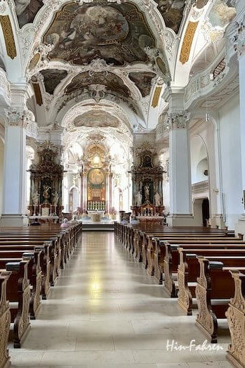Innenansicht eines Kirchenschiffs in der Nähe der Hohenzollernstraße, gesäumt von Holzbänken, die auf einen reich verzierten Altar unter einer gewölbten, bemalten Decke ausgerichtet sind.