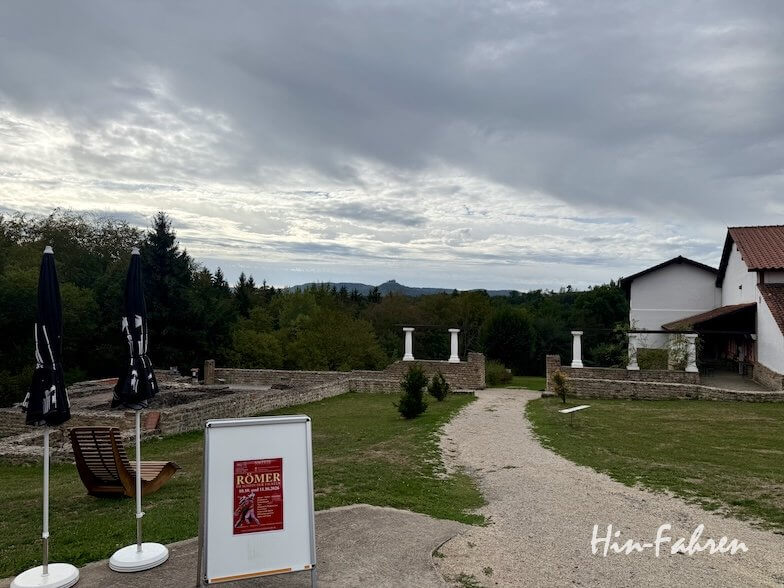 Ein Freilichtmuseum an der Hohenzollernstraße mit einem Schotterweg, römischen Ruinen, weißen Säulen und einem Veranstaltungsschild, das auf eine Römer-Ausstellung hinweist.