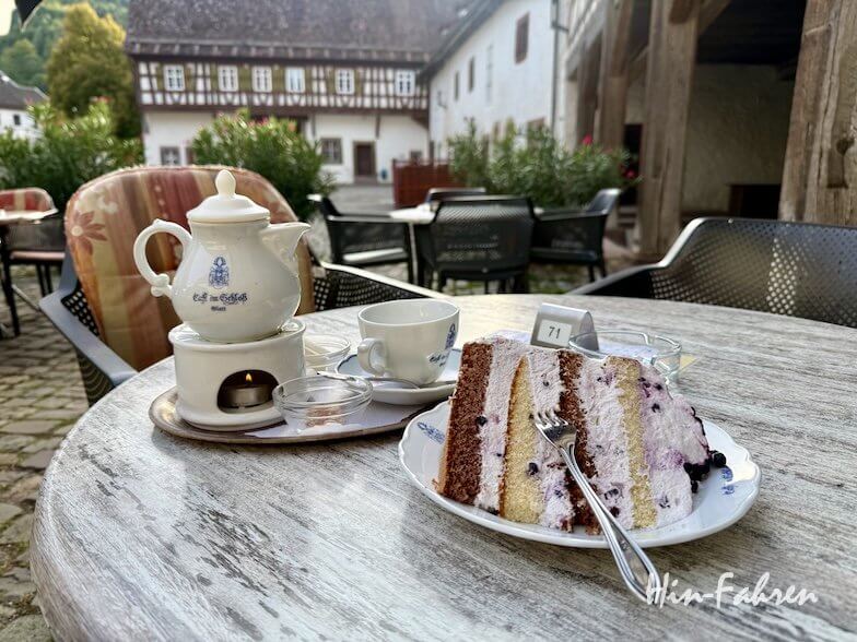 Ein Stück Blaubeer-Sahnetorte mit einer Gabel auf einem Teller neben einer Teekanne, die über einer Kerze auf einem Terrassentisch an der Hohenzollernstraße warm gehalten wird.