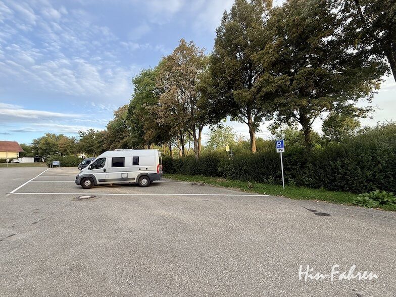Ein silbernes Wohnmobil der Marke Hohenzollernstraße stand auf einem asphaltierten Parkplatz neben einer Baumreihe und einem Parkschild.