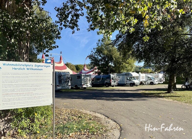 Vor einem Stellplatz, auf dem zahlreiche Wohnmobile stehen und im Hintergrund ein Zirkuszelt zu sehen ist, steht ein Schild, das Besucher auf dem Wohnmobilstellplatz in der Hohenzollernstraße in Sigmaringen willkommen heißt.